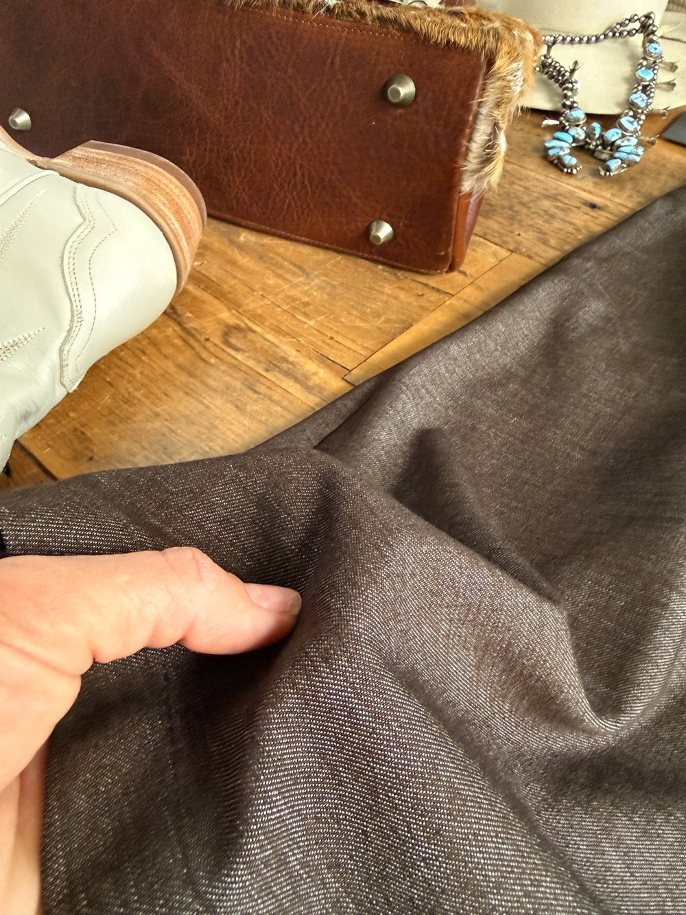 A hand pinches brown fabric on a wooden surface near a white boot, a leather bag with fur trim and metal studs, and a turquoise necklace—suggesting the finishing touches to an ensemble that could feature the Hazelwood Espresso Sleeveless Dress by Savannah Sevens western life{&}style.