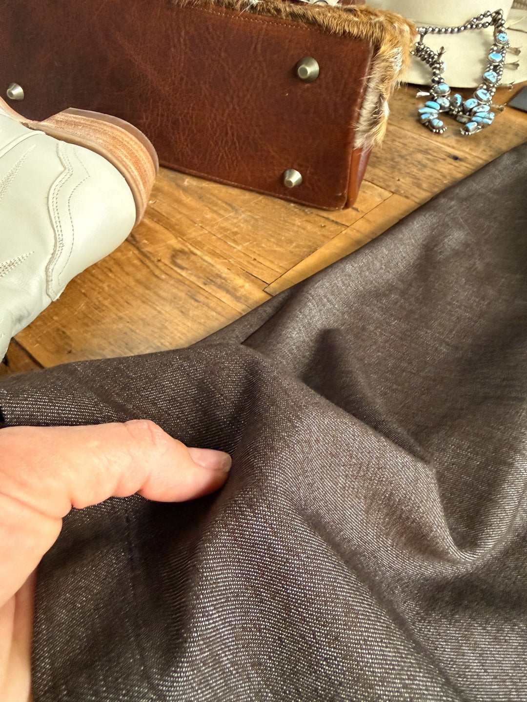 A hand pinches brown fabric on a wooden surface near a white boot, a leather bag with fur trim and metal studs, and a turquoise necklace—suggesting the finishing touches to an ensemble that could feature the Hazelwood Espresso Sleeveless Dress by Savannah Sevens western life{&}style.