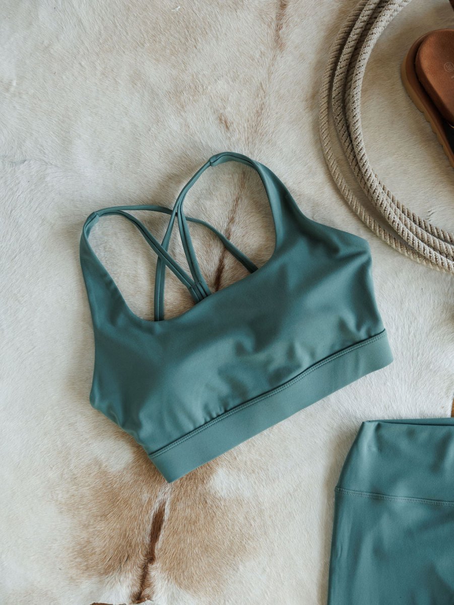 The Hazen Sports Bra {S} by Savannah Sevens western life{&}style, featuring cross back straps and compression fabric in green, is displayed on tan and white fur beside a coiled rope and a matching green garment.