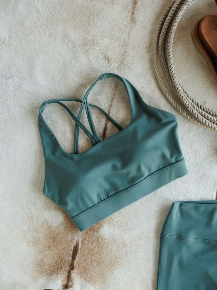 The Hazen Sports Bra {S} by Savannah Sevens western life{&}style, featuring cross back straps and compression fabric in green, is displayed on tan and white fur beside a coiled rope and a matching green garment.