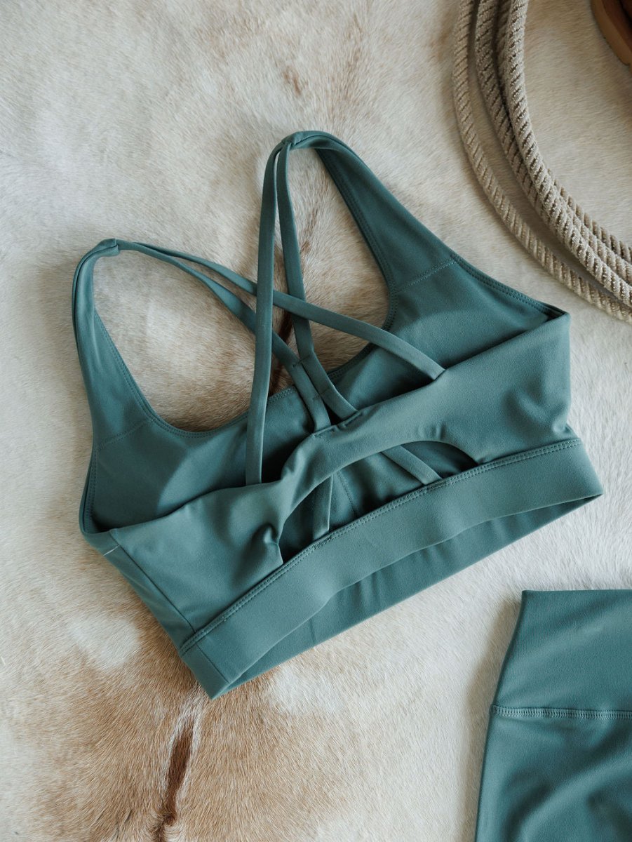The Hazen Sports Bra {S} by Savannah Sevens western life{&}style in green features cross back straps and supportive compression fabric. Displayed on a light animal hide with matching leggings and a coiled beige rope in the upper right corner.