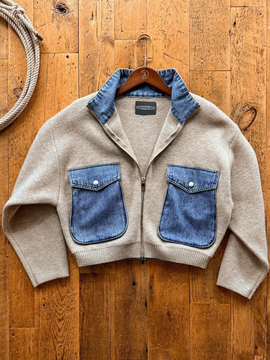 Hendley Oatmeal Knit Jacket in oatmeal-colored knit with a full zipper, featuring vintage wash denim accents on the collar and two front pockets, hanging on a wooden hanger against a wood floor.