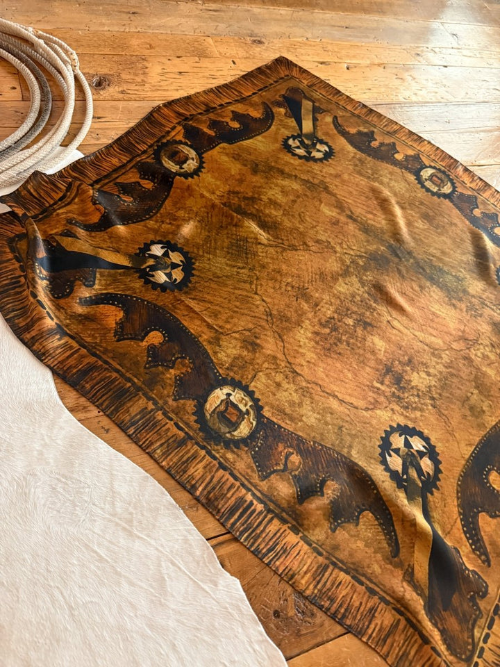 The Henryetta Wild Rag by Savannah Sevens western life{&}style, in brown and gold with black designs and conchos, is displayed on a wooden floor beside a white hide and golden bronze scarf.