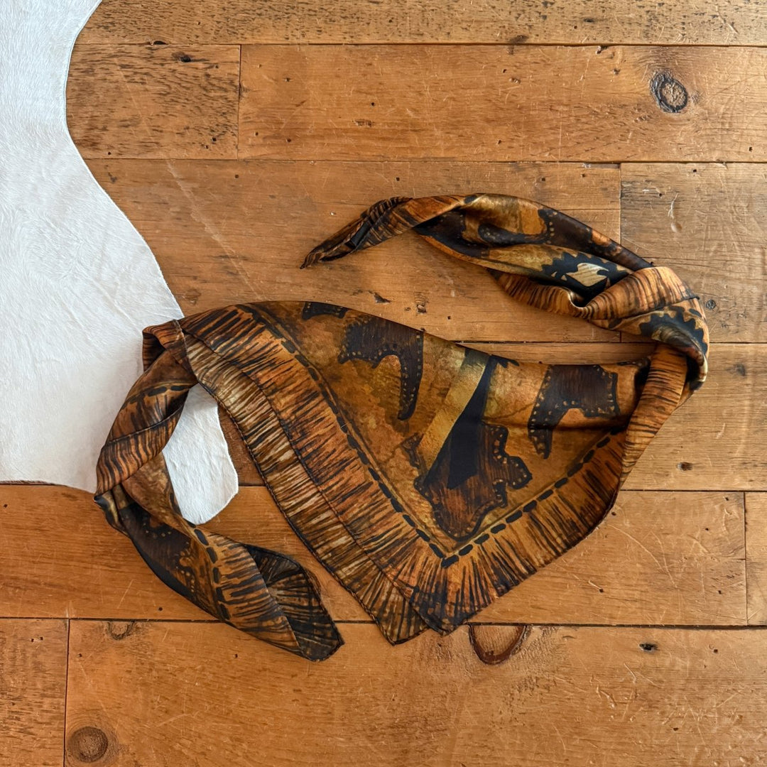The Henryetta Wild Rag by Savannah Sevens western life{&}style, featuring a brown pattern with tan and black accents, is styled like a western silk bandana and displayed on wood beside white cowhide.