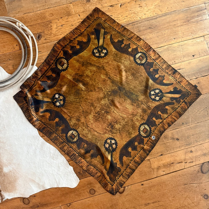 The Henryetta Wild Rag by Savannah Sevens western life{&}style, featuring brown and black western patterns, is displayed on a wooden floor beside a white animal hide rug and a golden bronze scarf.