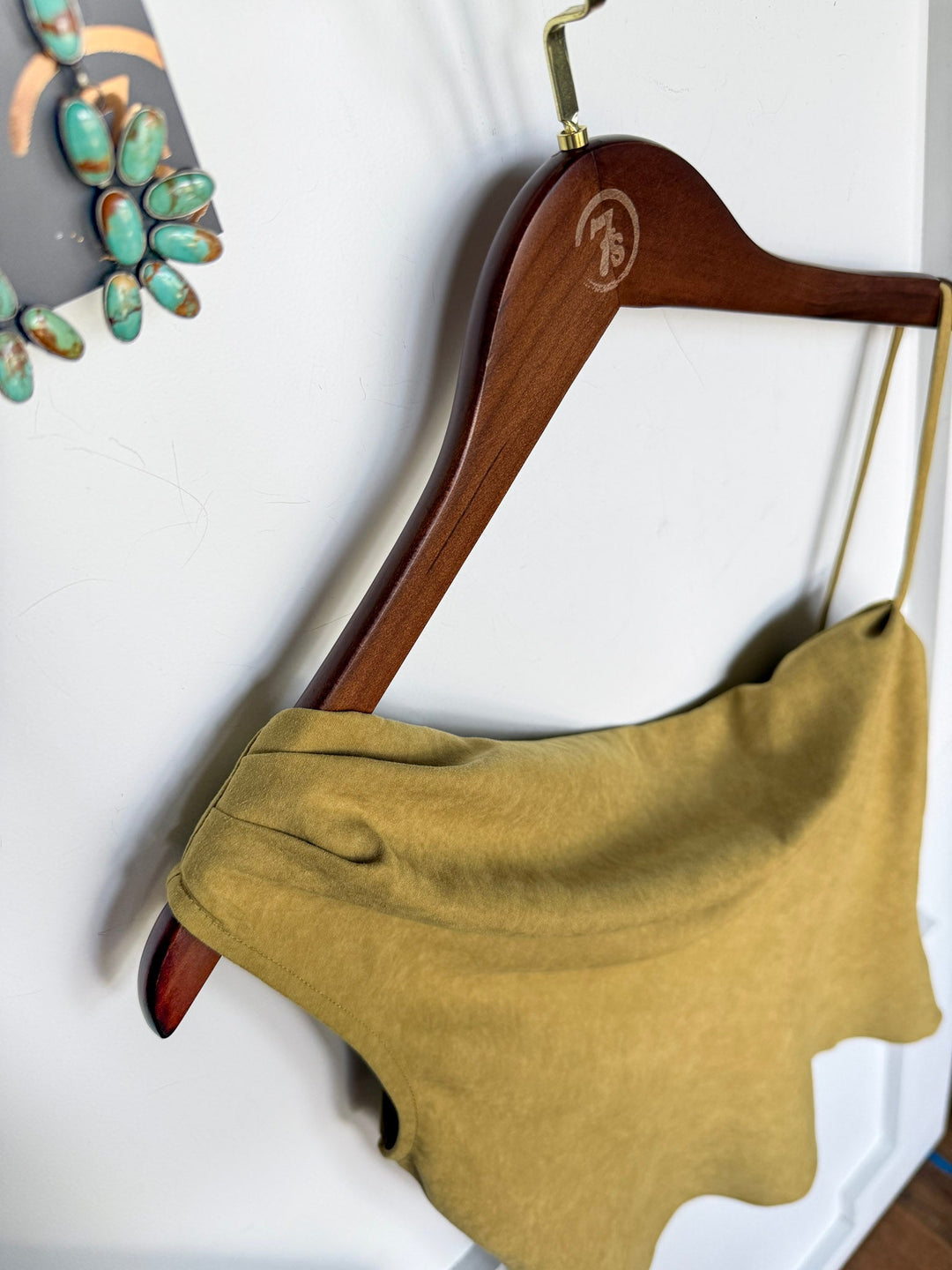 A wooden hanger displays the Hillard Chartreuse Sleeveless Top by Savannah Sevens western life{&}style—a light olive-green asymmetrical cowl neck top—against a white wall. Part of a turquoise and silver accessory appears along the left edge of the image.