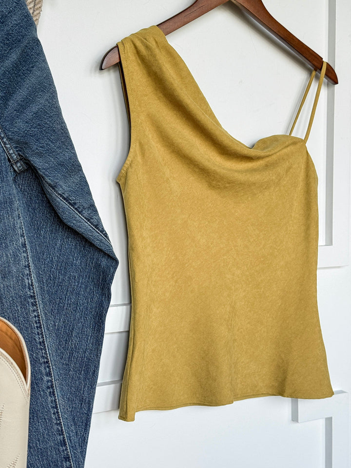 The Hillard Chartreuse Sleeveless Top by Savannah Sevens western life{&}style, featuring a yellow asymmetrical cowl neck design, hangs on a wooden hanger against a white wall. Blue jeans and a beige bag can be seen partially to the left.