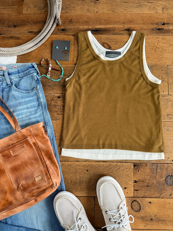 Flat lay of a casual outfit: blue jeans, brown leather bag, beaded bracelet, sunglasses, the Hillsboro Olive Layered Tank by Savannah Sevens western life{&}style in young contemporary sizing, white knit sneakers made from ultra soft cotton, coiled rope, and a square pin on a wooden floor.