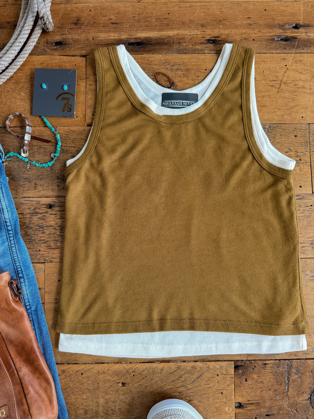 A Hillsboro Olive Layered Tank by Savannah Sevens western life{&}style, featuring young contemporary sizing for the perfect fit, is laid flat on a wooden surface layered over a white tank top, with a brown bag, denim, bracelet, rope, and a small card partially visible around it.