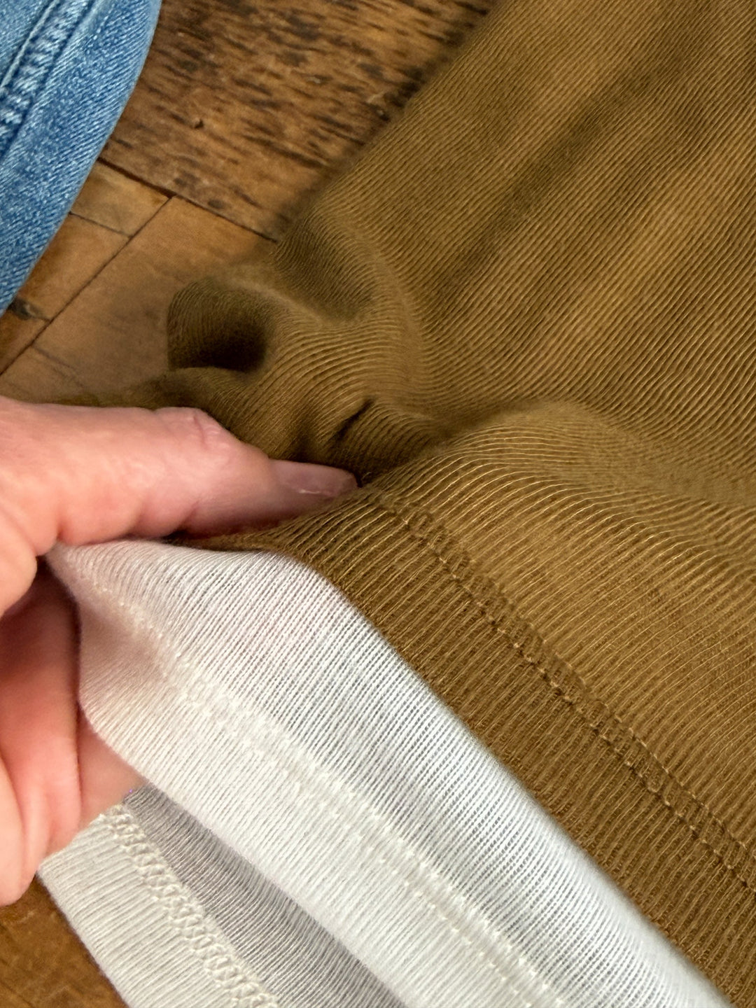 A hand holds the edge of a brown pant leg, revealing a sewn-on white fabric cuff. In the background, part of the person’s blue jeans and a wooden floor are visible, providing contrast to the ultra-soft cotton look of the Savannah Sevens western life{&}style Hillsboro Olive Layered Tank—perfect for young contemporary sizing.