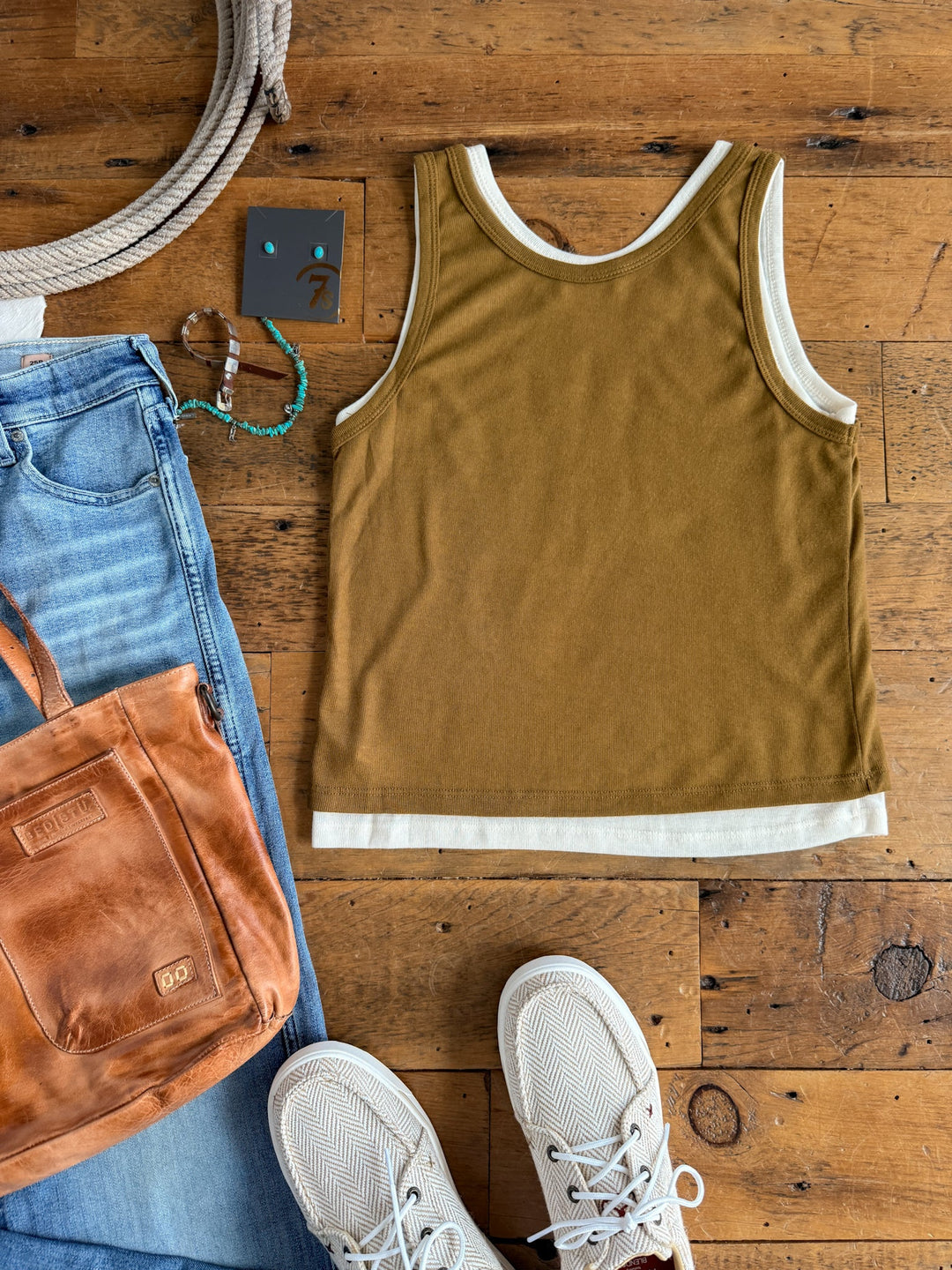Flat lay of a casual outfit featuring the Hillsboro Olive Layered Tank by Savannah Sevens western life{&}style in ultra soft cotton over a cream tank, paired with light blue jeans, white sneakers, a brown leather crossbody bag, turquoise bracelet, gray earrings, and a coiled rope—all arranged on a wooden floor.
