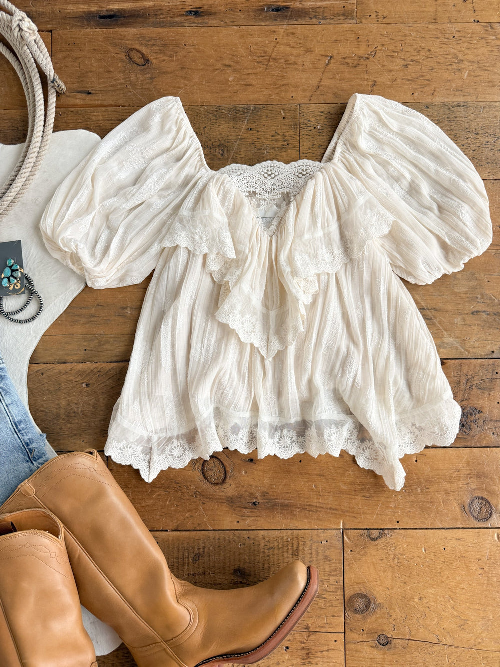 The Holbrook Cream Lace Trim Top by Savannah Sevens western life{&}style is displayed on a wooden floor, with tan cowboy boots, blue jeans, and turquoise jewelry partially visible nearby.