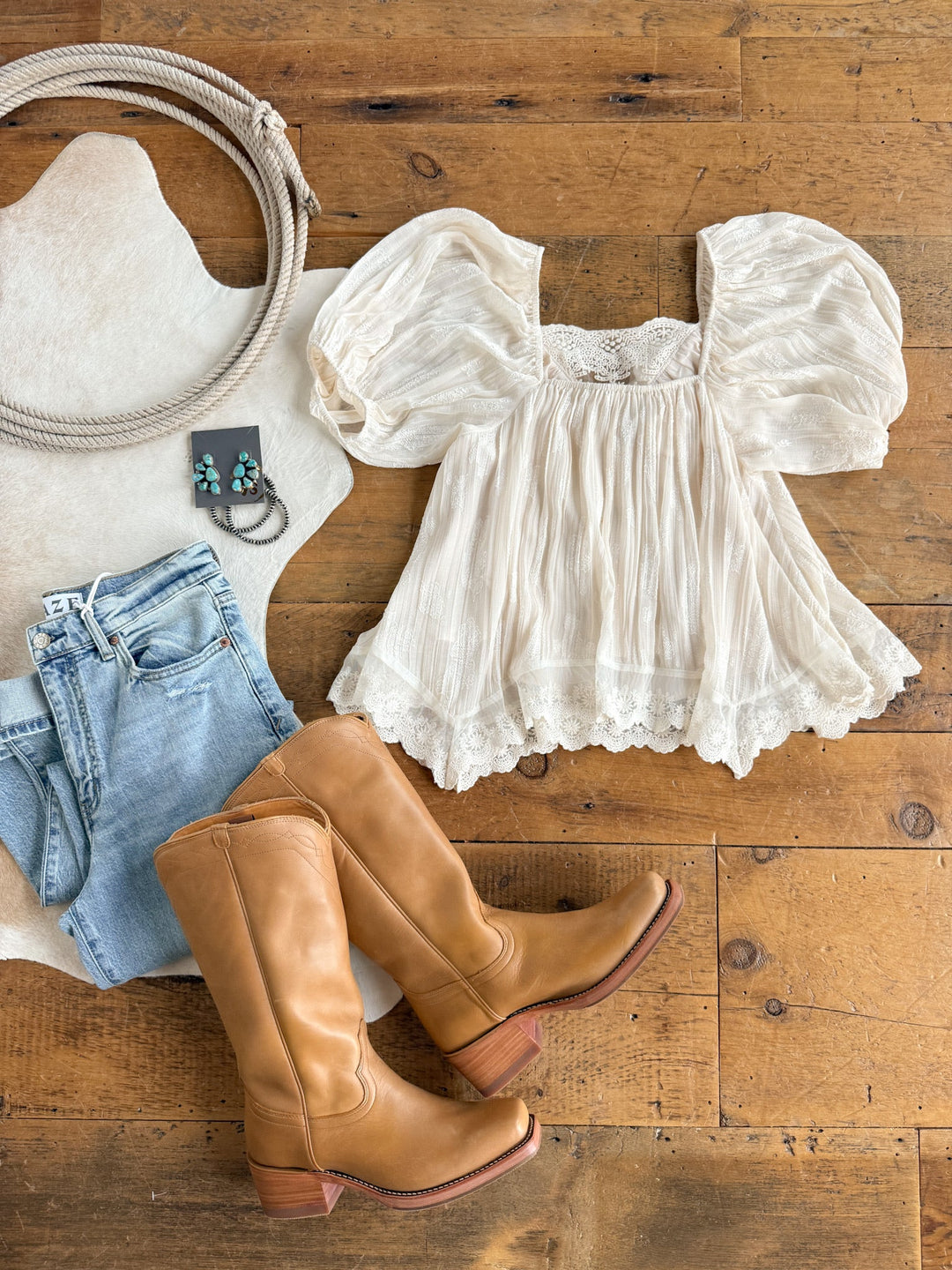 Flat lay of the Holbrook Cream Lace Trim Top by Savannah Sevens western life{&}style, paired with light blue jeans, tan knee-high boots, a coiled rope, turquoise earrings, and a white cowhide rug on a wooden floor.