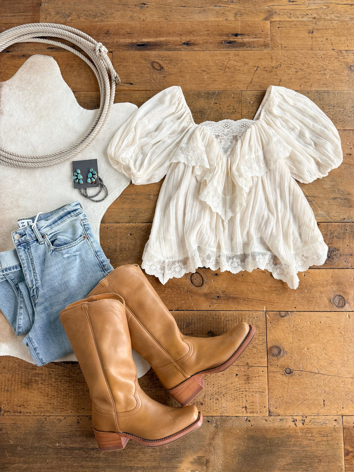 The Holbrook Cream Lace Trim Top by Savannah Sevens western life{&}style is styled with blue jeans, tan knee-high boots, and a turquoise pendant necklace, all arranged on a wooden floor with a white cowhide rug.