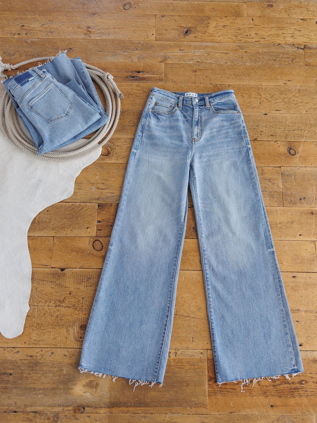 The Holland High Rise Wide Leg Jean by Savannah Sevens western life{&}style in a medium light wash with frayed hems is laid flat on a wooden floor, while another folded pair sits on a cream cowhide rug.