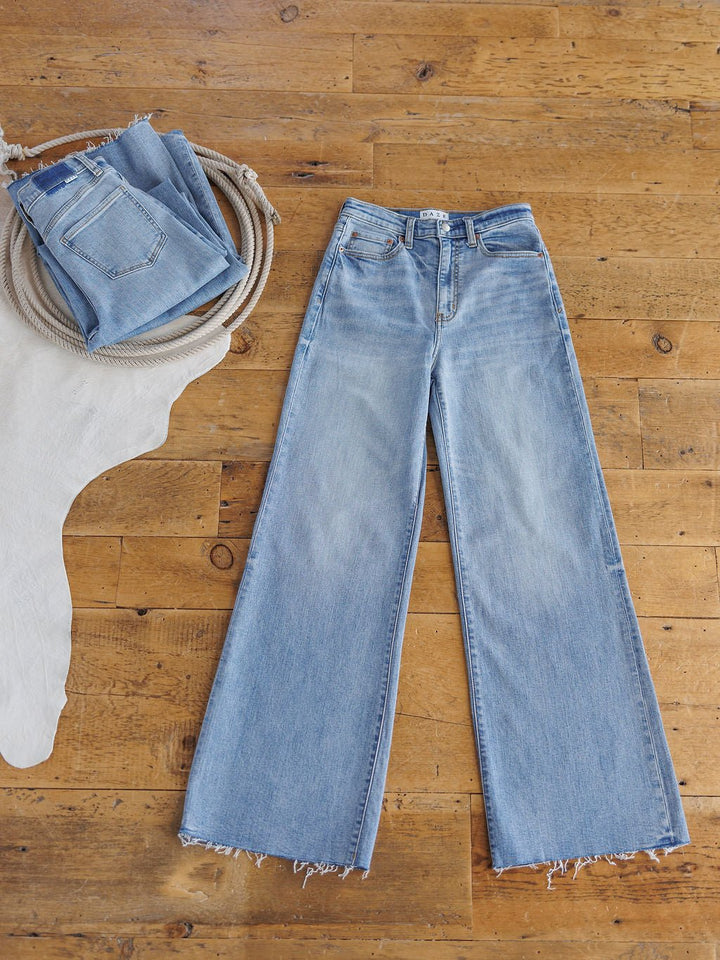 The Holland High Rise Wide Leg Jean by Savannah Sevens western life{&}style in a medium light wash with frayed hems is laid flat on a wooden floor, while another folded pair sits on a cream cowhide rug.