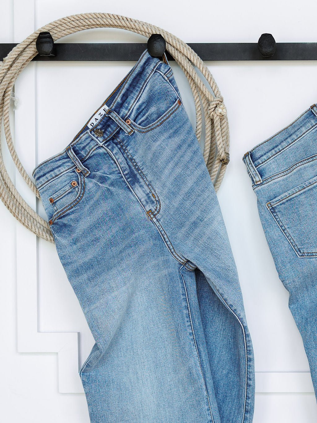 The Holland High Rise Wide Leg Jean by Savannah Sevens western life{&}style in medium light wash hangs on a black wall hook with a coiled rope, while another pair of wide leg jeans is partially visible on the right.