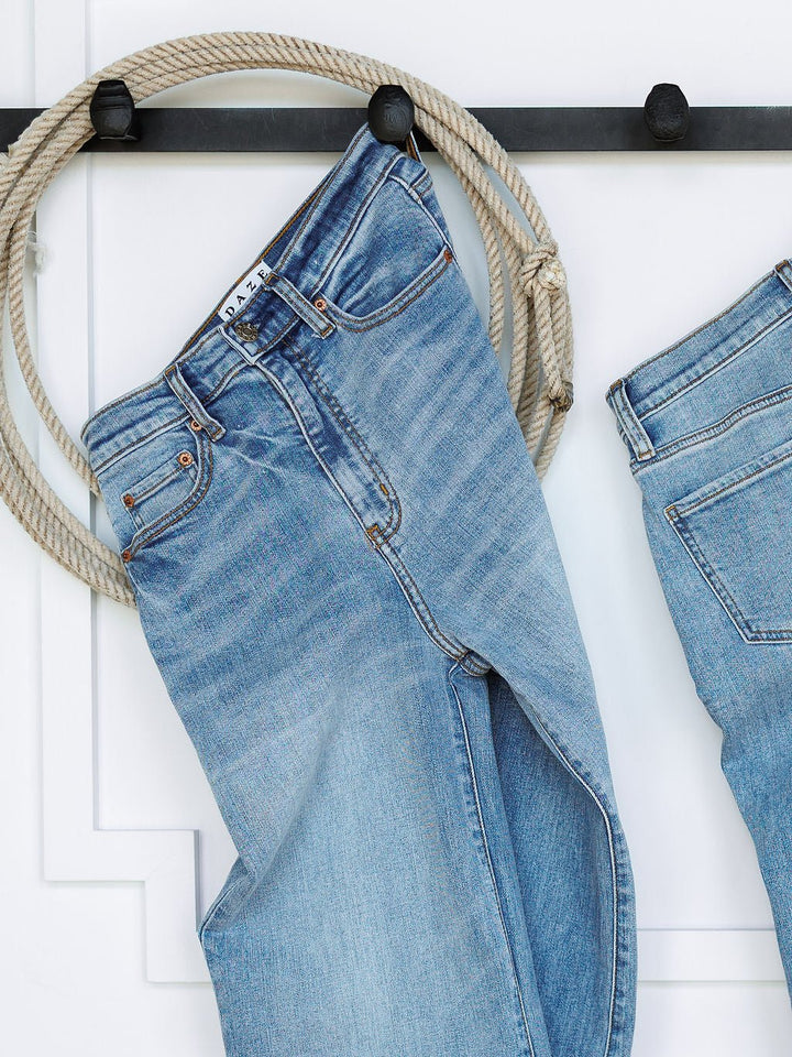 The Holland High Rise Wide Leg Jean by Savannah Sevens western life{&}style in medium light wash hangs on a black wall hook with a coiled rope, while another pair of wide leg jeans is partially visible on the right.