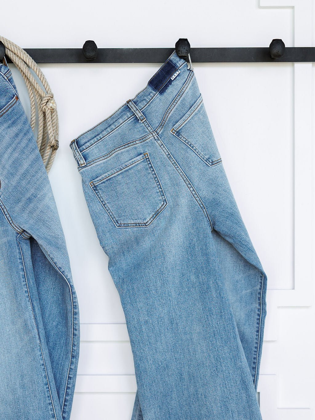 A pair of medium light wash The Holland High Rise Wide Leg Jeans by Savannah Sevens western life{&}style hang on a black wall hook against a white background, with part of another ultra high rise 90's wide leg jean visible on the left.