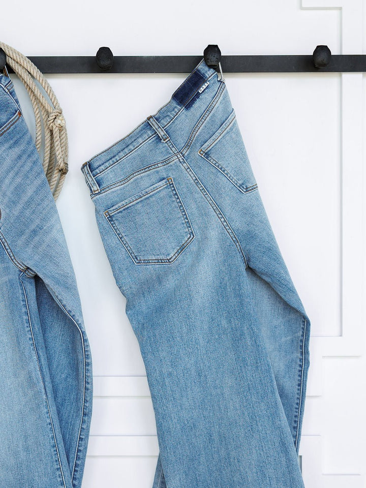 A pair of medium light wash The Holland High Rise Wide Leg Jeans by Savannah Sevens western life{&}style hang on a black wall hook against a white background, with part of another ultra high rise 90's wide leg jean visible on the left.