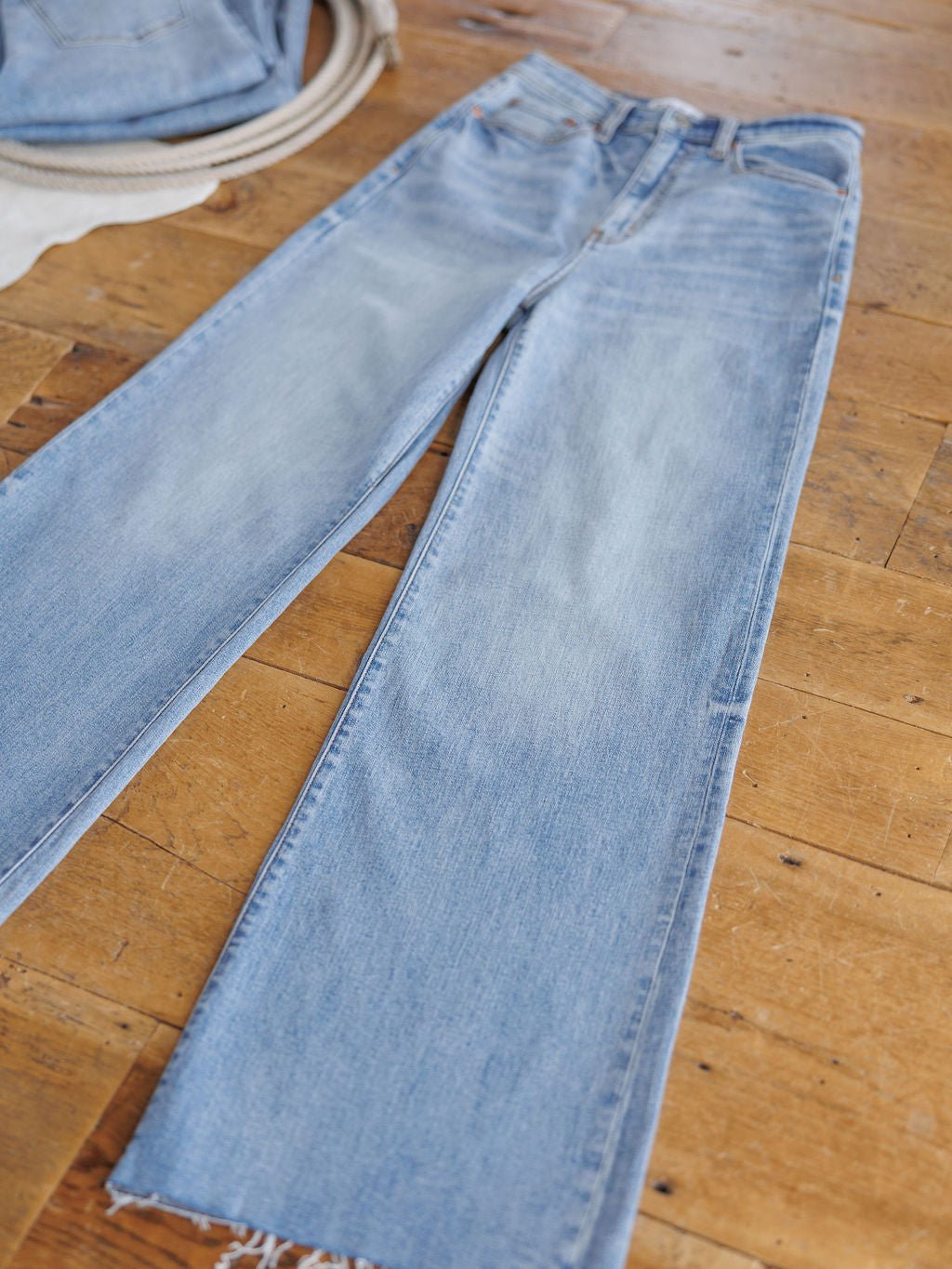 The Holland High Rise Wide Leg Jean by Savannah Sevens western life{&}style features a medium light wash, straight-leg fit, raw frayed hems, and an ultra high rise channeling '90s vibes. Laid flat on wood floor with a beige garment in the top left corner.