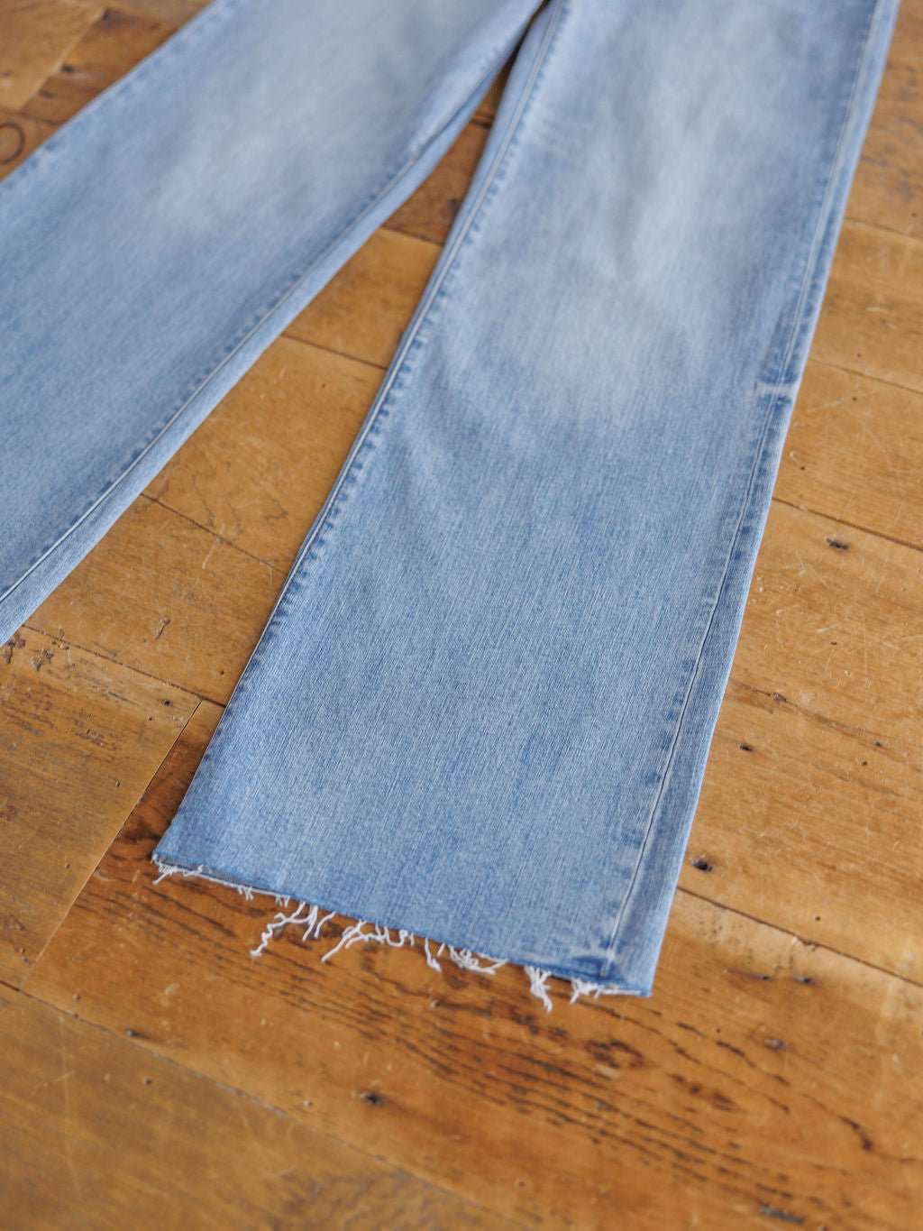 The Holland High Rise Wide Leg Jean by Savannah Sevens western life{&}style, featuring a medium light wash and frayed hems, is shown lying flat on a wooden floor.
