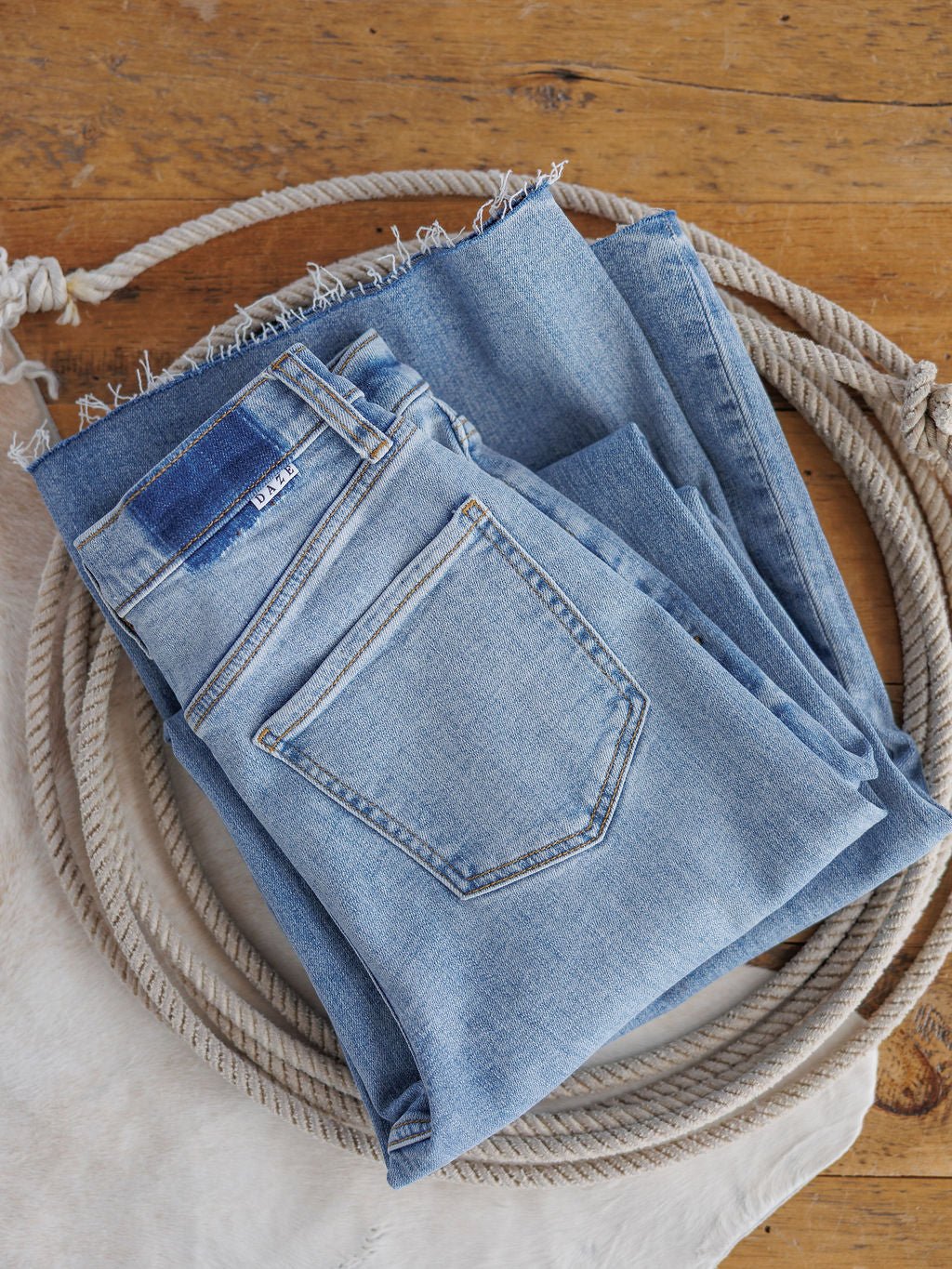 The Holland High Rise Wide Leg Jean by Savannah Sevens western life{&}style in a medium light wash features a 90's wide leg fit and frayed hem, shown folded atop a coiled rope on a wooden surface with light fabric underneath.