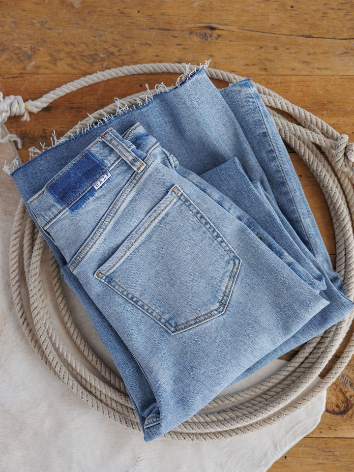 The Holland High Rise Wide Leg Jean by Savannah Sevens western life{&}style in a medium light wash features a 90's wide leg fit and frayed hem, shown folded atop a coiled rope on a wooden surface with light fabric underneath.