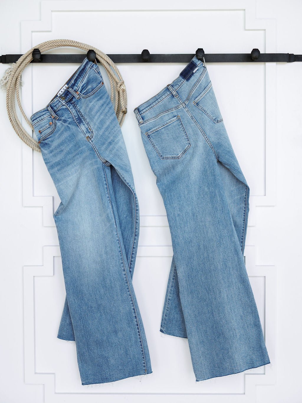 Two pairs of The Holland High Rise Wide Leg Jean by Savannah Sevens western life{&}style in medium light wash hang side by side on a black wall rack with a coiled rope on the left, set against a white wall with decorative molding.