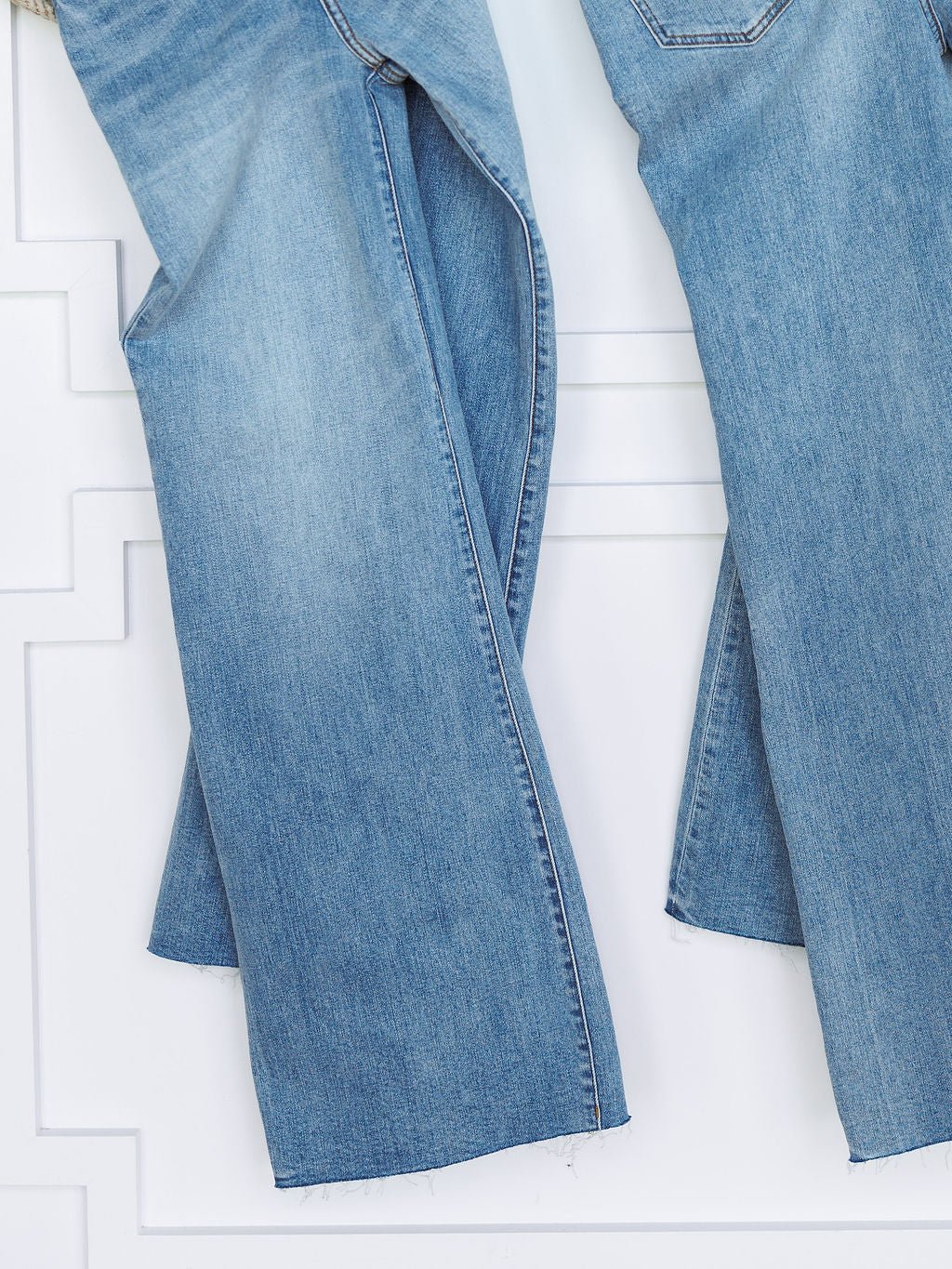 A close-up of two pairs of The Holland High Rise Wide Leg Jean by Savannah Sevens western life{&}style in medium light wash denim with frayed hems, hanging side by side against a white textured wall.