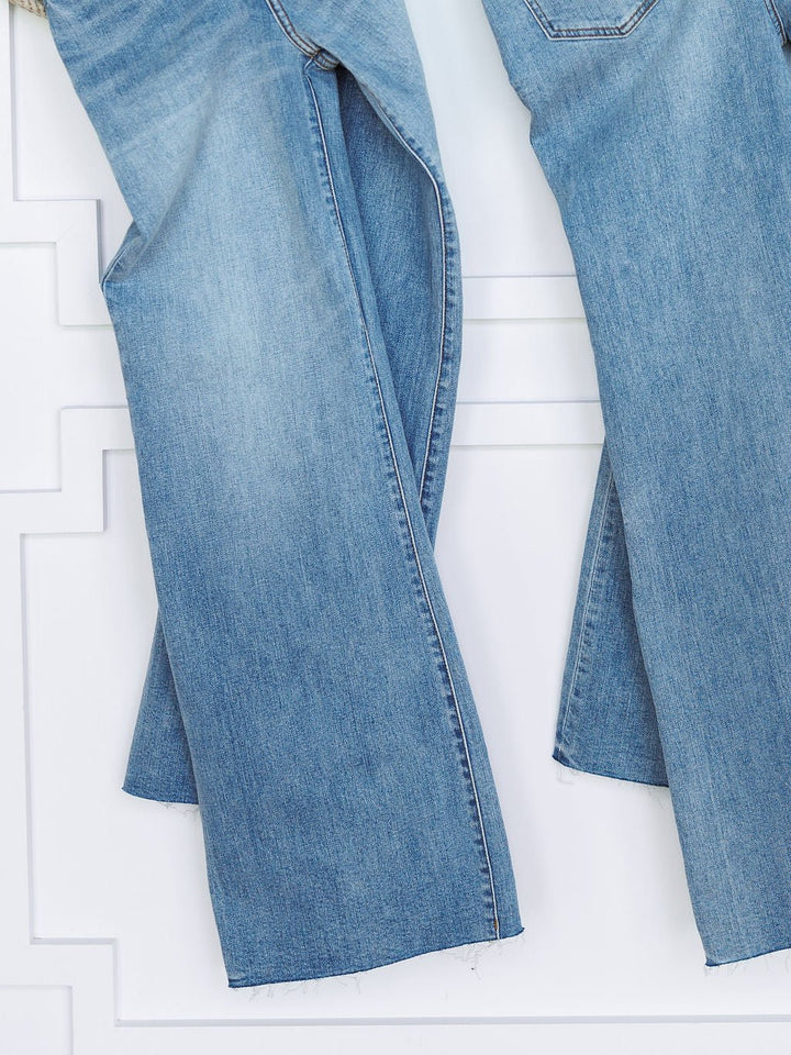A close-up of two pairs of The Holland High Rise Wide Leg Jean by Savannah Sevens western life{&}style in medium light wash denim with frayed hems, hanging side by side against a white textured wall.