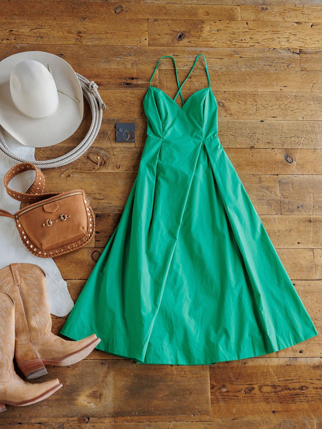 The Hollandale Jade Green Dress by Savannah Sevens western life{&}style, featuring petite adjustable straps, is styled on a wooden floor with tan cowboy boots, a tan hat, a studded handbag, and silver hoop earrings.