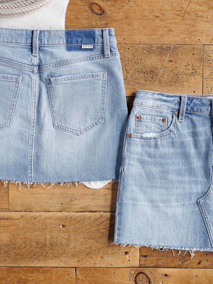 Two The Hollyn Skirts by Savannah Sevens western life{&}style in medium light wash denim with raw cut hems are laid flat on a wooden floor, one showing the back pockets and the other displaying front details.