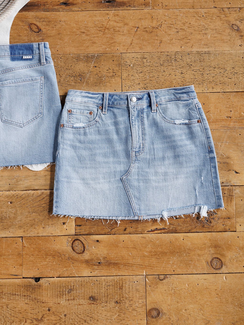 The Hollyn Skirt by Savannah Sevens western life{&}style, a distressed medium-light wash denim skirt with a raw cut hem, is displayed flat on a wooden floor beside another denim piece.