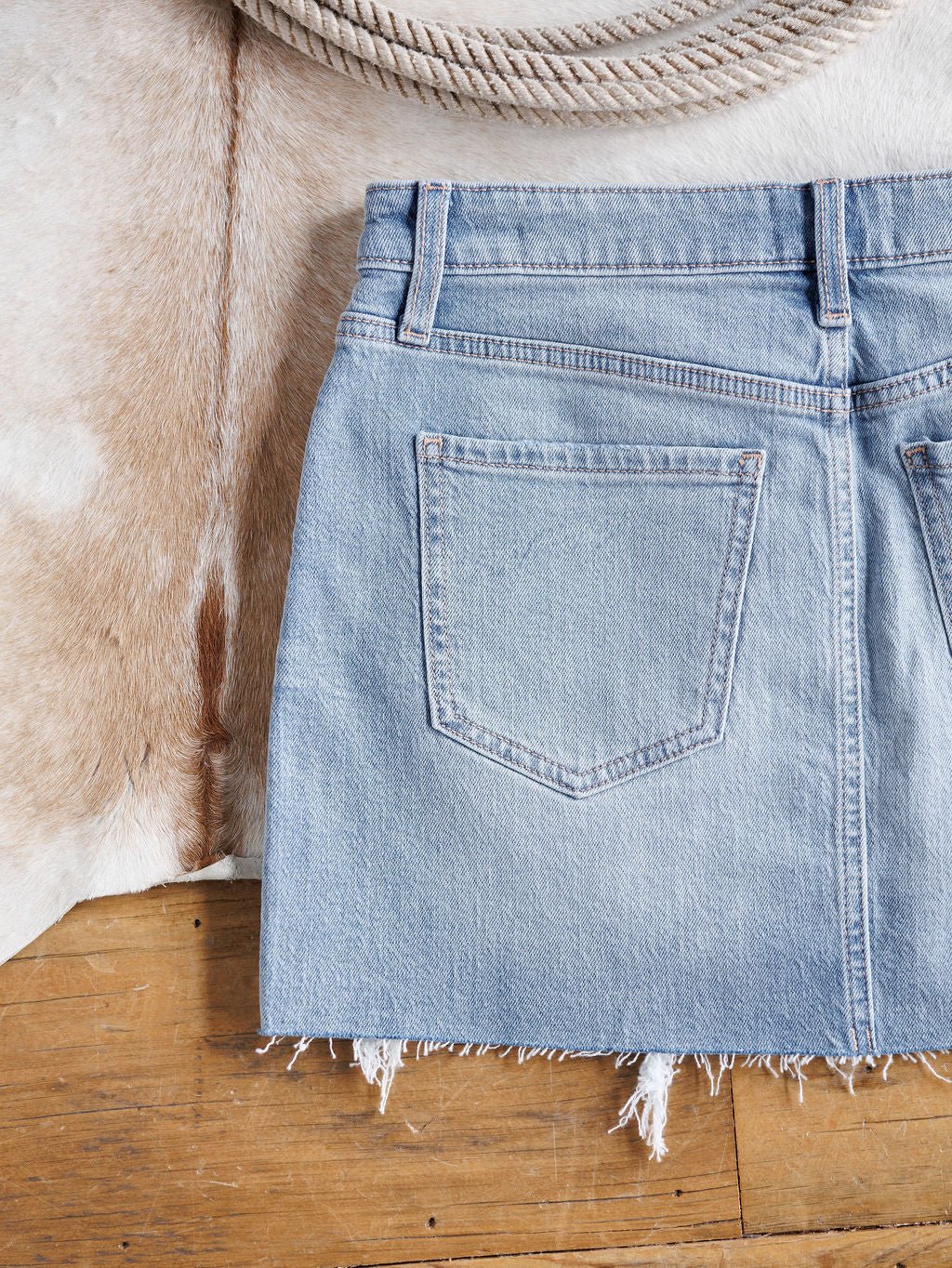 The Hollyn Skirt by Savannah Sevens western life{&}style is a medium light wash distressed denim skirt with a raw cut hem, shown from the back with one pocket, placed on a wooden floor partially over a cowhide rug and coiled rope.