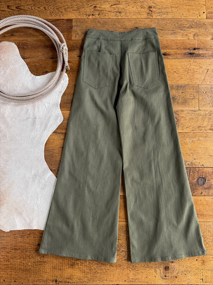 The Holt Olive Wide Leg Pants by Savannah Sevens western life{&}style, featuring a high-waisted fit and back pockets, are displayed on a wooden floor beside a white cowhide rug.