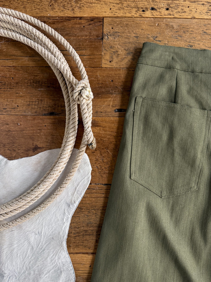 The Holt Olive Wide Leg Pants by Savannah Sevens western life{&}style are displayed on a wooden surface alongside a coiled rope and a piece of white cowhide, showcasing the pants’ distinctive back pocket.