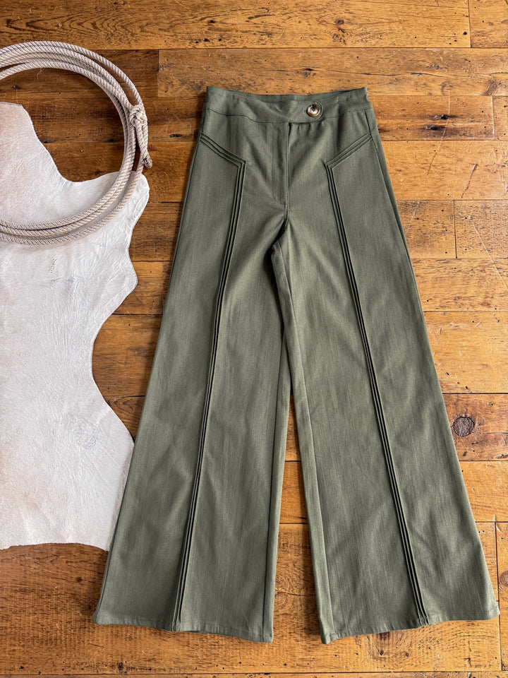 The Holt Olive Wide Leg Pants by Savannah Sevens western life{&}style, featuring a high waisted fit with pintuck front seam details and a button waist, are laid flat on a wooden floor beside a coil of rope and a piece of white leather.