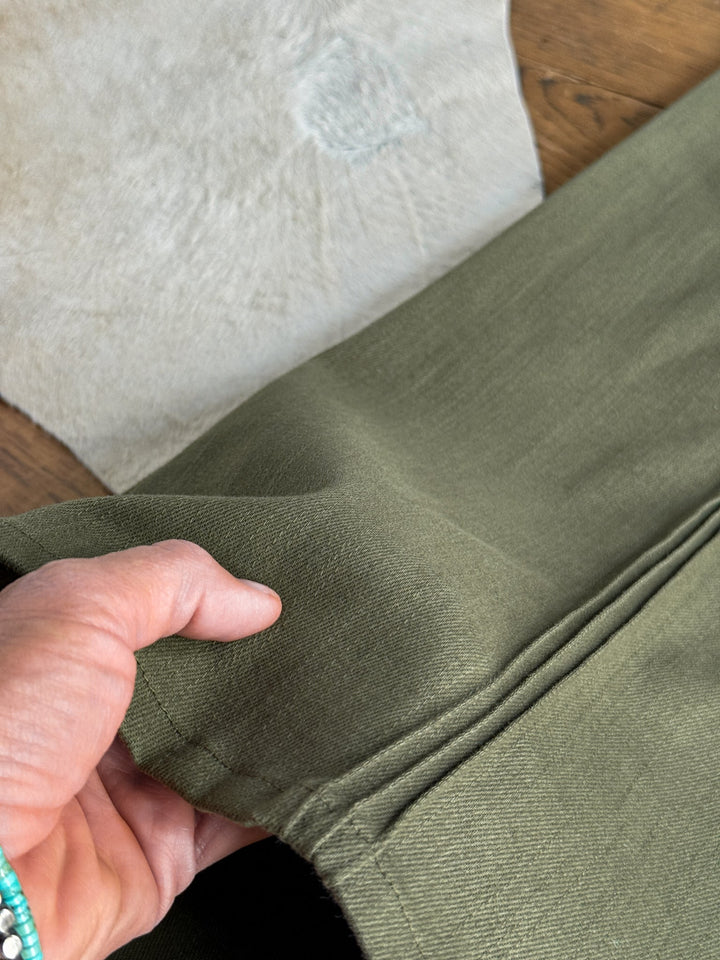 A hand holds the hem of the Holt Olive Wide Leg Pants by Savannah Sevens western life{&}style, showcasing the fabric and front pintuck seam details. A light-colored textured background and a wooden surface complete the setting.