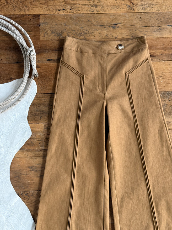 The Holt Saddle Tan Wide Leg Pants from Savannah Sevens western life{&}style, featuring stitched vertical seams and a single metallic button, are displayed on a wooden floor. To the left, a neatly coiled rope and a white textured object add rustic accents to the scene.