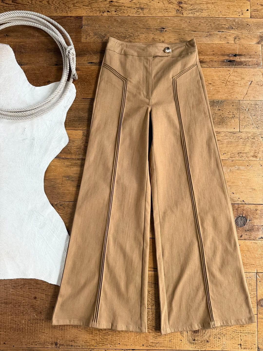 The Holt Saddle Tan Wide Leg Pants by Savannah Sevens western life{&}style, featuring high waisted triple pintuck seam detailing, are laid flat on a wooden floor. To the left, a coiled rope and a partially visible piece of white textured material complete the scene.