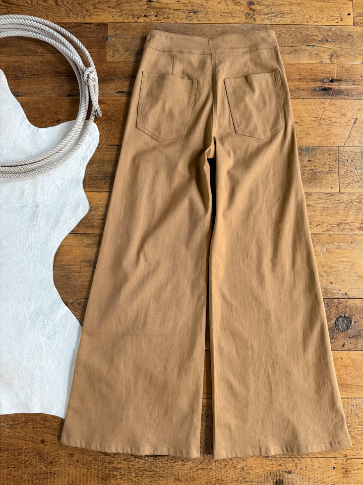 The Holt Saddle Tan Wide Leg Pants from Savannah Sevens western life{&}style are laid flat on a rustic wooden floor, accompanied by a coiled rope and a piece of white textured fabric. Designed with back pockets and a triple pintuck seam for added style and structure, these high-waisted pants feature a wide-leg silhouette in classic saddle tan.