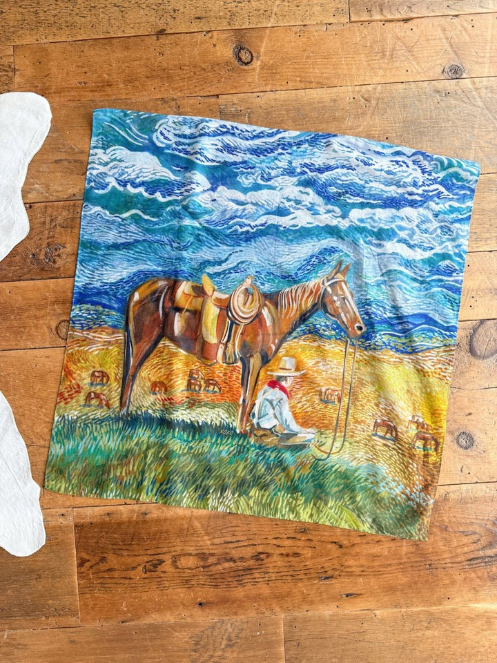 The Home On The Range Neck Scarf by Savannah Sevens western life{&}style features a cowboy beside his saddled horse with a swirling sky and grazing cattle, displayed on a wooden floor.