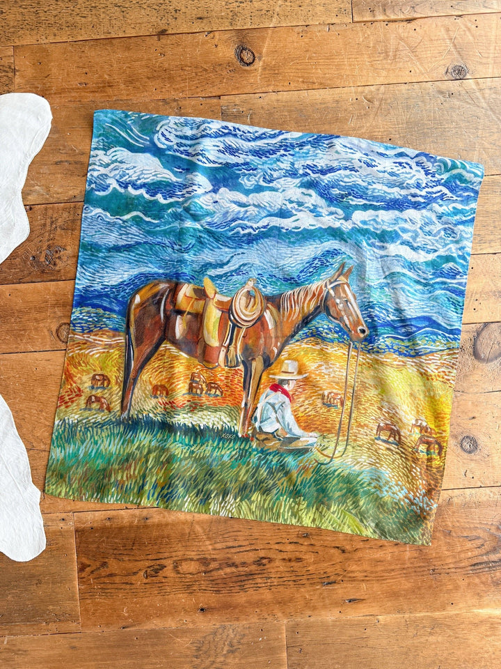 The Home On The Range Neck Scarf by Savannah Sevens western life{&}style features a cowboy beside his saddled horse with a swirling sky and grazing cattle, displayed on a wooden floor.
