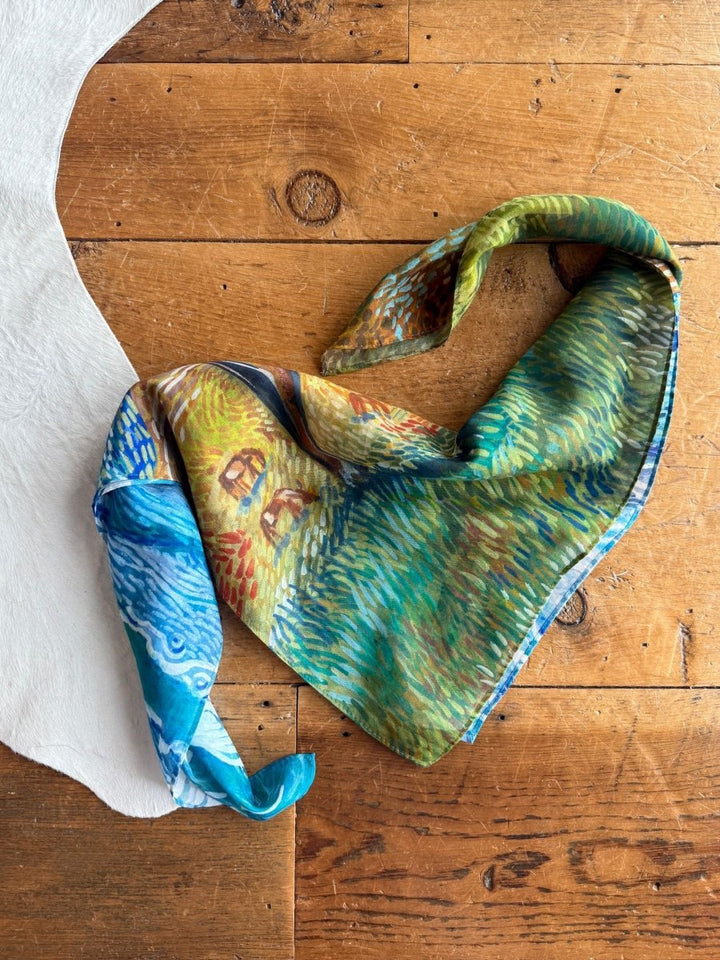 The Home On The Range Neck Scarf by Savannah Sevens western life{&}style, featuring a Van Gogh-inspired design, is arranged in a heart shape on rustic wood beside white fabric.