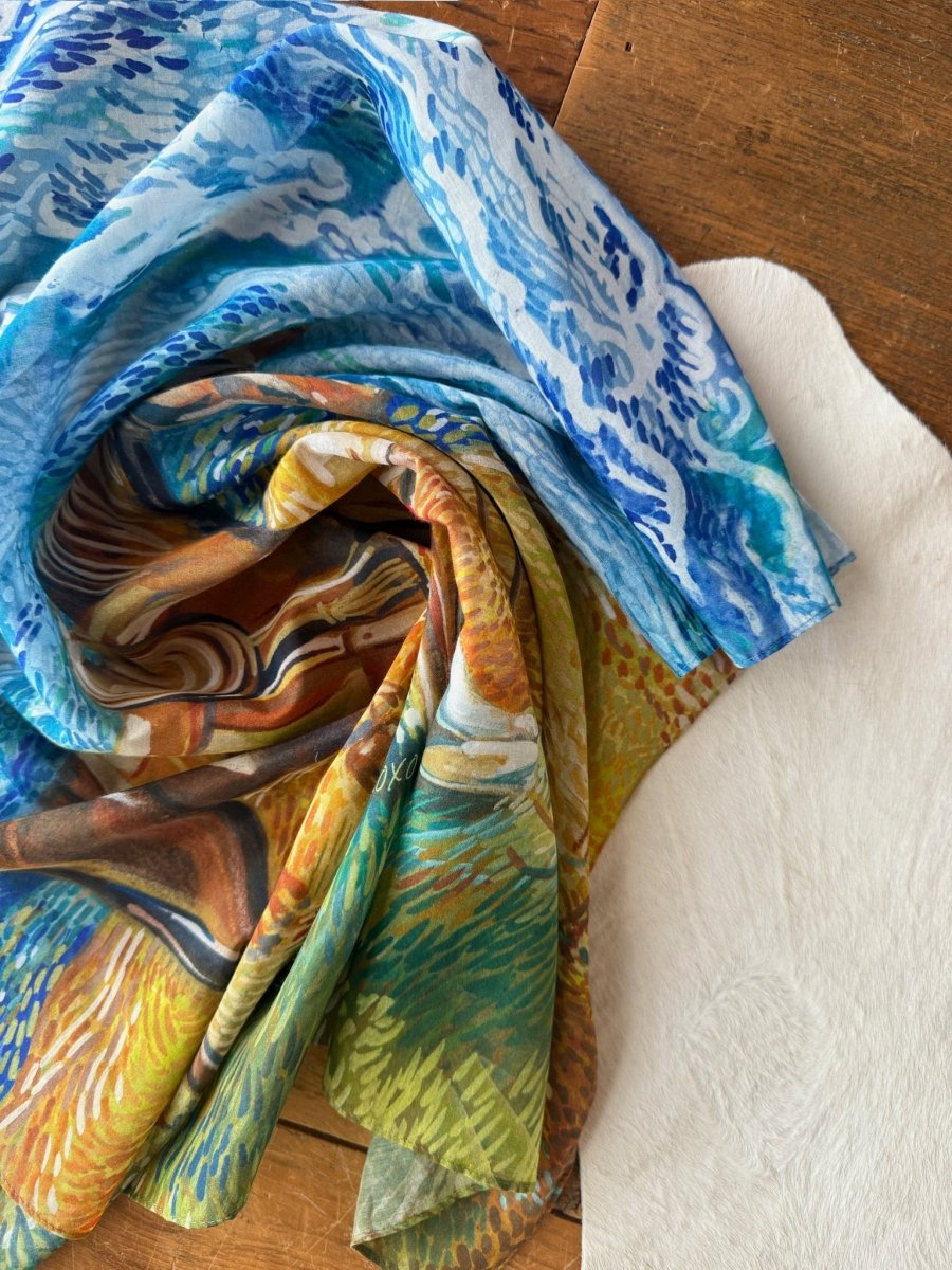 The Home On The Range Neck Scarf by Savannah Sevens western life{&}style, featuring blue, green, and brown swirls, is draped over a wooden surface beside white textured fabric.