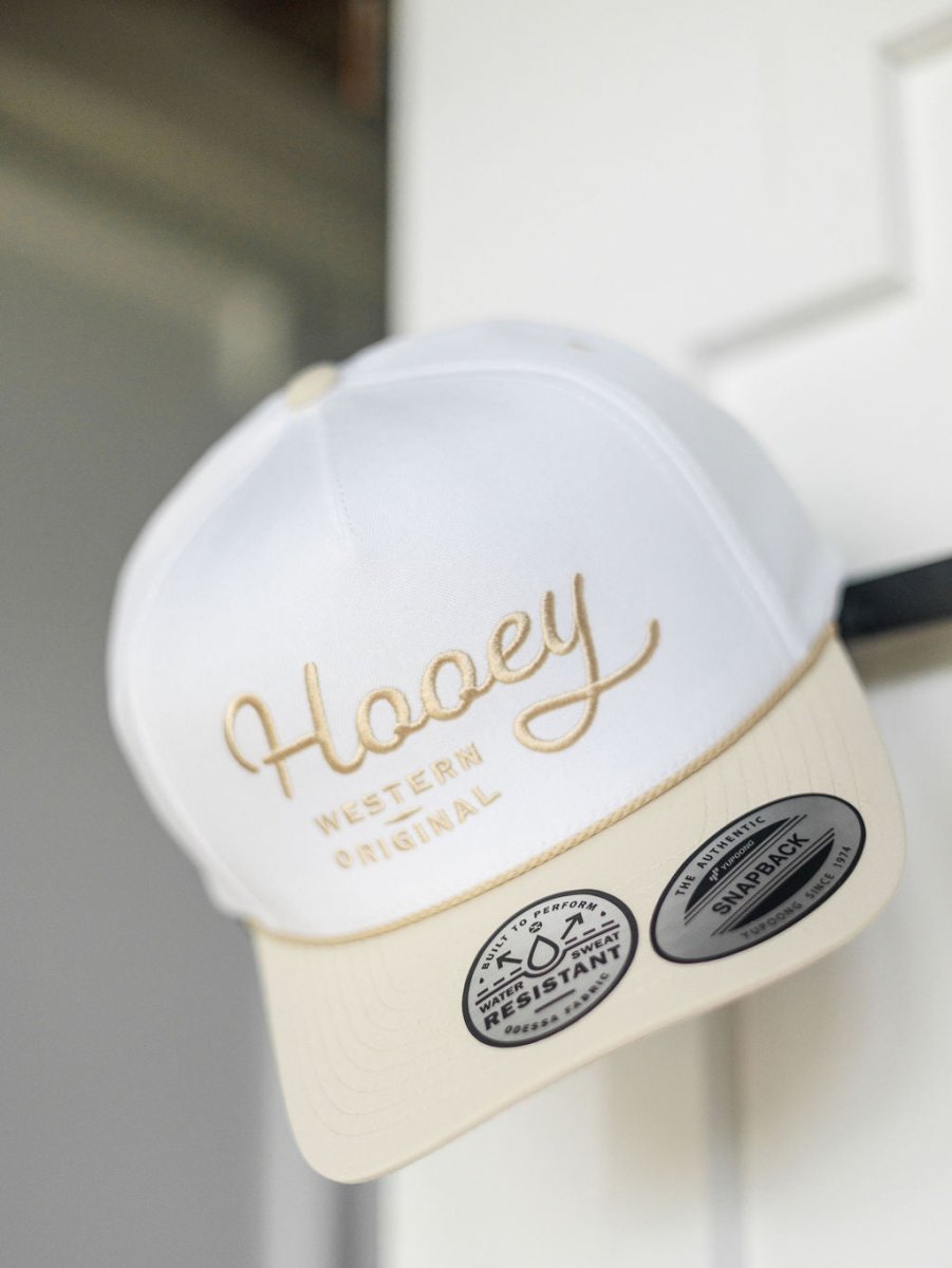 A white and beige Hooey Benton Cap by Savannah Sevens western life{&}style hangs on a door, showcasing "Western Original" embroidered on the front and two stickers on its adjustable snapback brim.