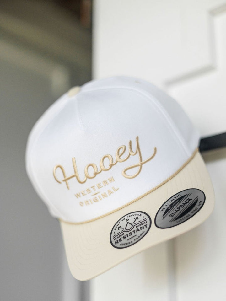A white and beige Hooey Benton Cap by Savannah Sevens western life{&}style hangs on a door, showcasing "Western Original" embroidered on the front and two stickers on its adjustable snapback brim.