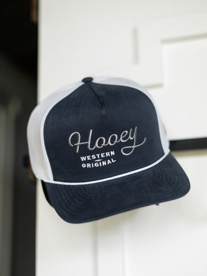 A navy blue and white Hooey Colby Cap by Savannah Sevens western life{&}style with "Western Original" embroidery hangs on a white door, featuring an adjustable snap back for comfort.