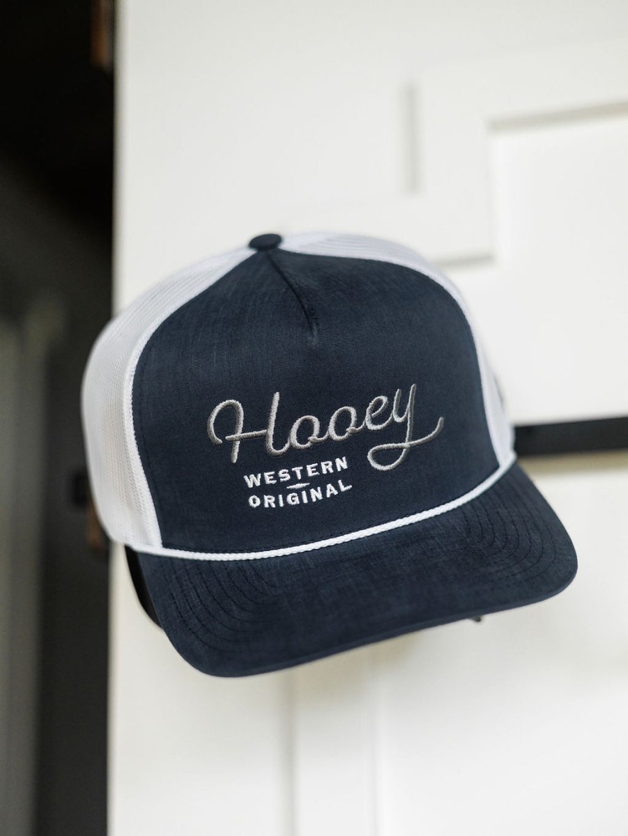 A navy blue and white Hooey Colby Cap by Savannah Sevens western life{&}style with "Western Original" embroidery hangs on a white door, featuring an adjustable snap back for comfort.